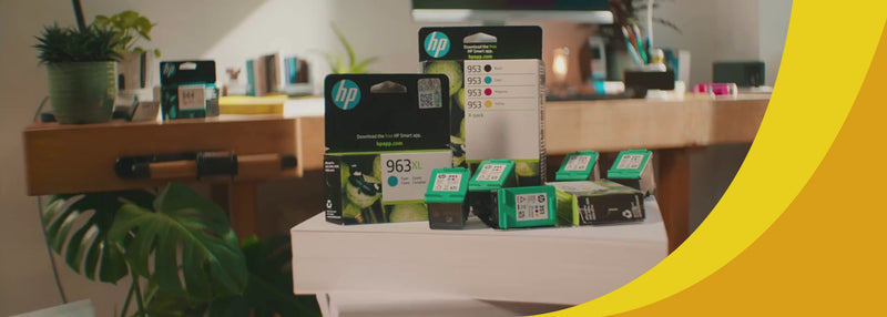 Im Vordergrund sieht man Schachteln von HP Druckerpatronen sowie einzelnen HP Druckerpoatrone, die auf einem Stapel Papier liegen. Im Hintergund sieht man eine Schreibtischumgebung.